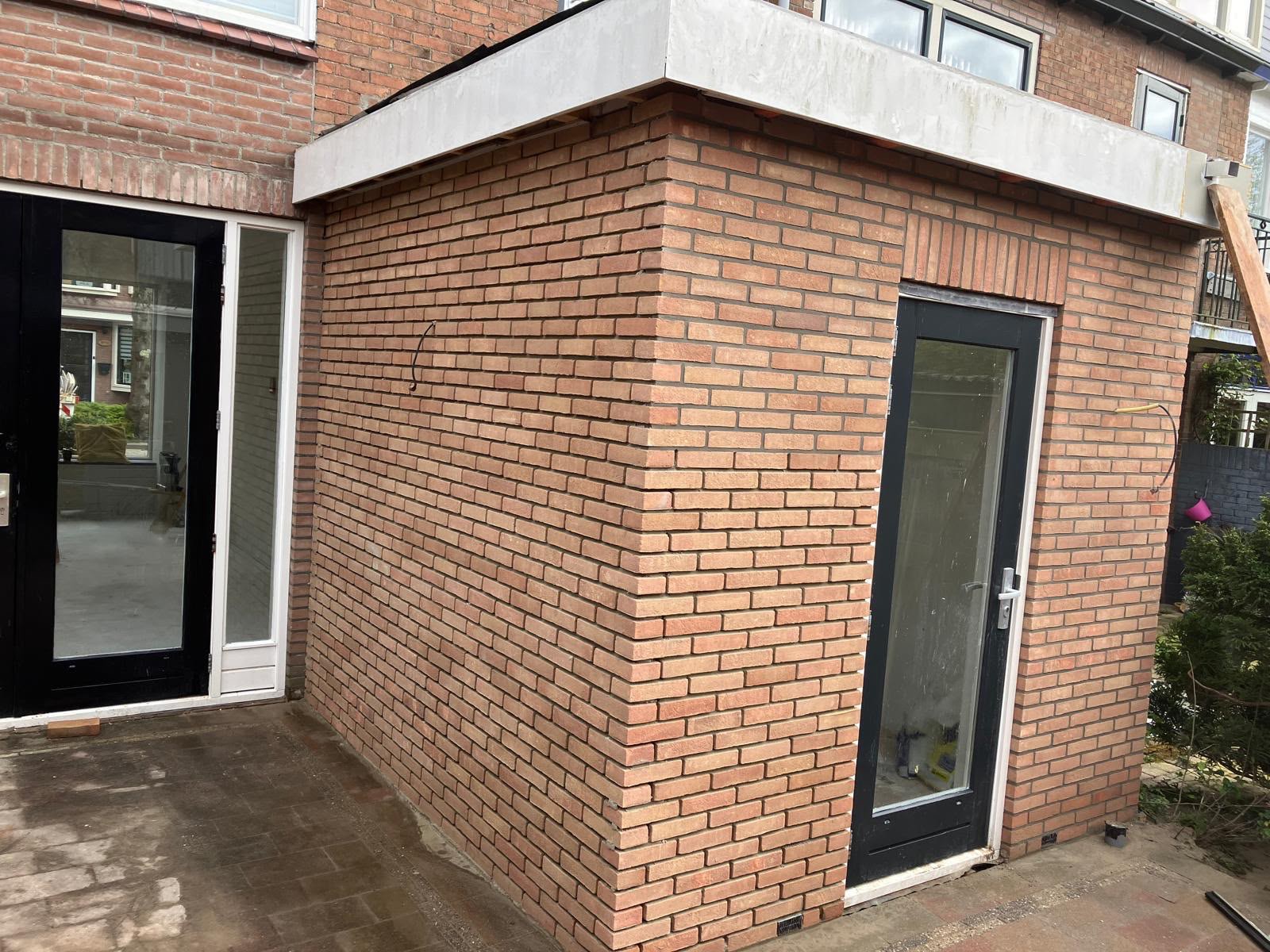 Gemetselde aanbouw met een zwarte deur, naast openslaande deuren